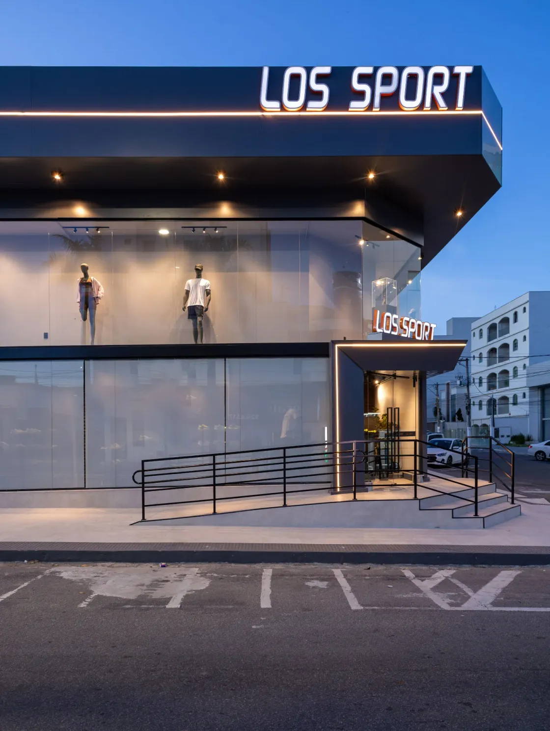 Entrada da loja Los Sport com marquise iluminada, rampa de acesso e vitrine de pé-direito duplo ao anoitecer