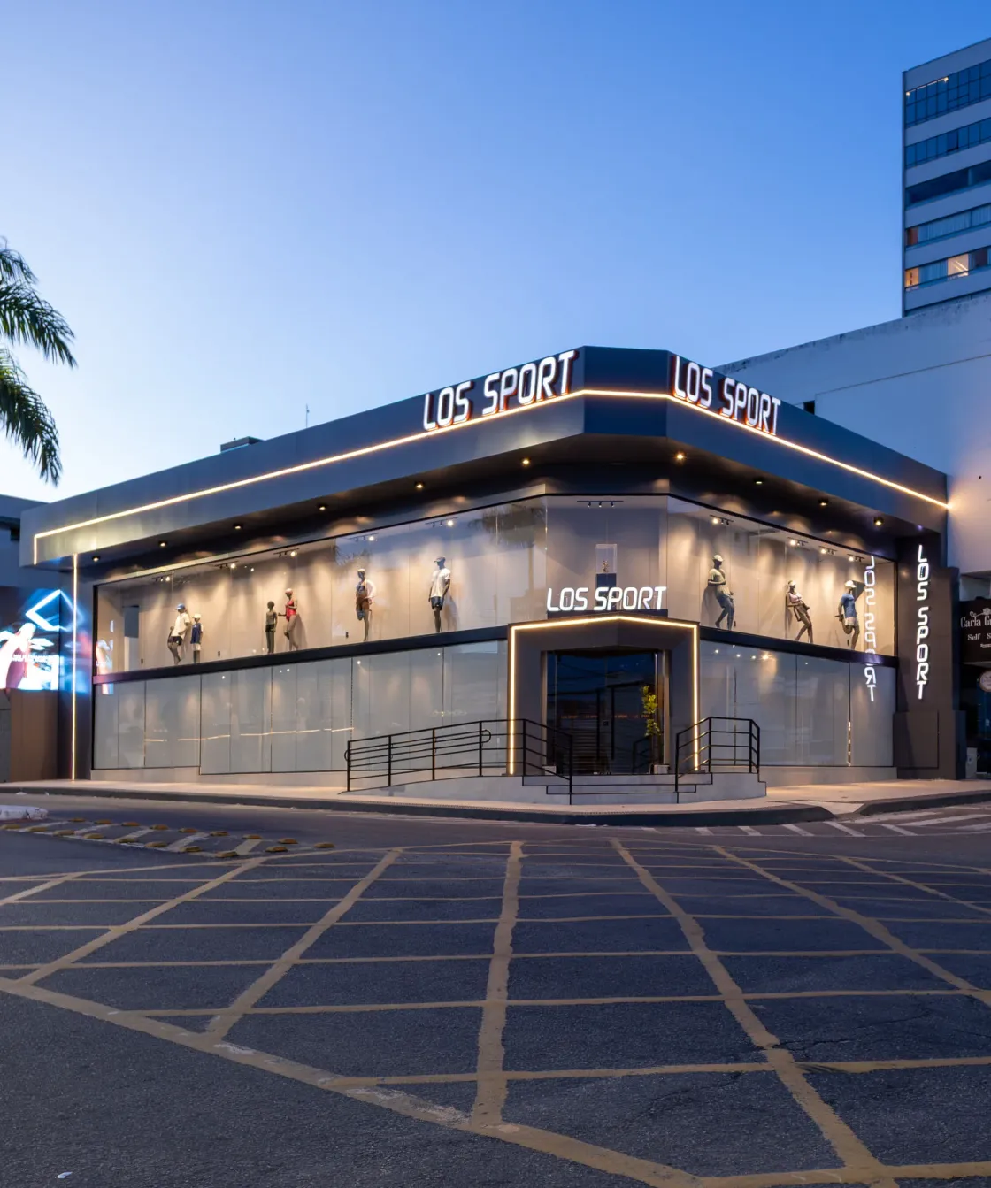 Fachada de esquina da loja Los Sport em painéis escuros, letreiro iluminado e faixa contínua de luz quente ao entardecer